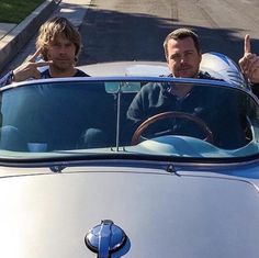 two men sitting in the drivers seat of a car giving thumbs up to someone else