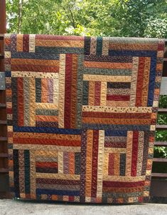 a multicolored quilt hanging on a wooden fence in front of some green trees