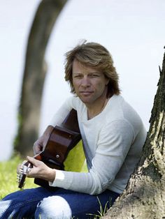 a man sitting in the grass with a guitar next to a tree and looking at his cell phone