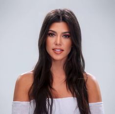 a woman with long dark hair wearing a white off the shoulder top and looking at the camera