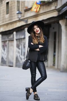 a woman in black is standing on the street with her arms crossed and wearing a hat