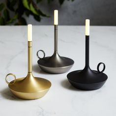 three different types of modern candlesticks on a marble countertop with plants in the background