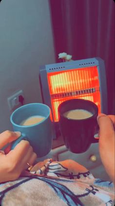 two people are toasting and drinking coffee in front of an electric heater on a bed