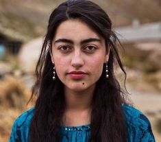 a woman with long hair and earrings standing in front of a dirt road, looking at the camera