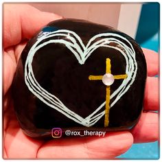 a hand holding a painted rock with a cross in the shape of a heart on it