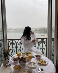 a woman sitting at a table with food and drinks in front of an open window