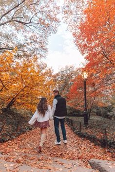 a man and woman holding hands while walking through an autumn park with leaves on the ground