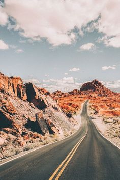 an empty road in the middle of nowhere with mountains and clouds behind it on a sunny day