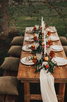 a long wooden table topped with white plates and place settings next to a tree filled with flowers