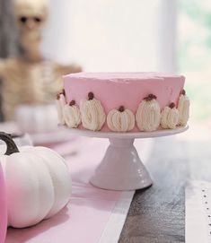 a pink frosted cake sitting on top of a table next to white pumpkins