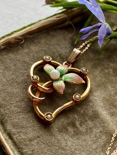 A 9CT gold Art Nouveau Style enamel floral heart pendant set with a border of seed pearls.:The hand painted enamel flower is in subtle shades of green and pink. The flower has a central Pearl. The scrolling border has all the whiplash  features of the Art Nouveau movement and softened with the addition of five small seed pearls. The piece has some age as the seed Pearl colour has darkened with age.  The bail is a simple wedge shaped one and the fine trace chain is modern and 9CT gold. The price Art Nouveau Gold Enamel Jewelry, Gold Enamel Art Nouveau Jewelry, Victorian Gold Enamel Necklace, Elegant Enamel Heart Pendant Necklace, Elegant Heart Pendant Enamel Necklace, Vintage Heart Enamel Necklaces, Vintage Enamel Heart Necklaces, Vintage Heart Enamel Necklace, Vintage Enamel Heart Necklace