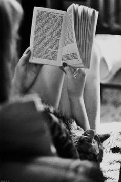 a woman reading a book while laying on her stomach with a cat sleeping next to her