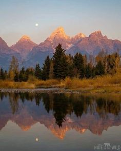 the mountains are reflected in the still water at sunset, with trees and grass around them