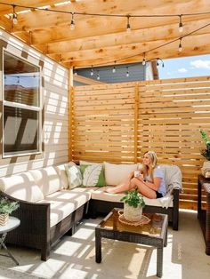 a woman sitting on top of a couch next to a wooden table under a pergolated roof