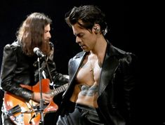 a man with no shirt on playing an electric guitar next to another man in leathers