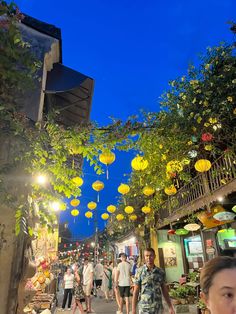 many people are walking down the street with yellow lanterns hanging from the buildings above them