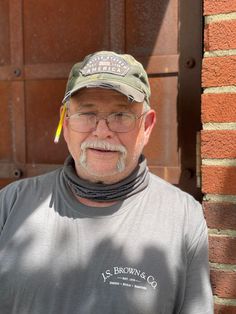 an older man with glasses and a hat standing in front of a brick wall wearing a gray t - shirt