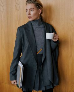 a woman leaning against a wall holding a coffee cup and folders in her hands