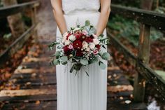 a woman in a white dress holding a red and white bouquet on a wooden bridge