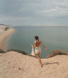 a woman in white dress walking on sand dunes next to the ocean with her arms outstretched