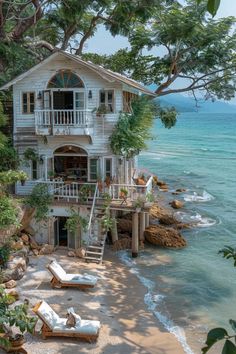 a house on the beach with lounge chairs in front of it and trees over looking the water