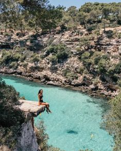 a woman sitting on the edge of a cliff next to a body of blue water