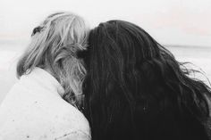 two women with long hair standing next to each other on the beach looking at the ocean