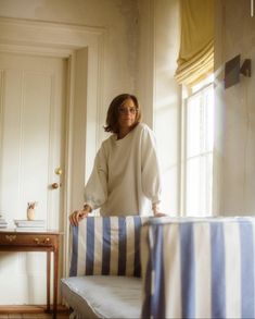 a woman standing on top of a bed next to a wooden table with a blue and white striped cover