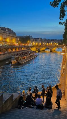 people are sitting on the side of a river watching boats go by in the distance