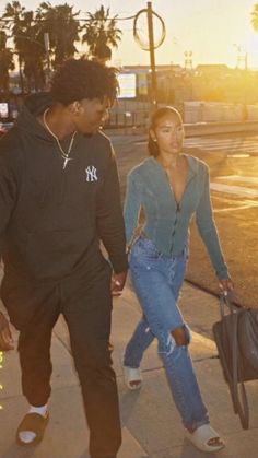 a man and woman walking down the street with luggage in hand while the sun is setting