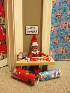 an elf is sitting at a table with some wrapping paper on it and there is a sign that says may's workshop