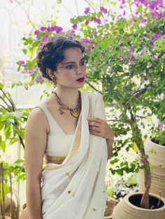 a woman wearing a white sari and gold jewelry standing next to a potted plant