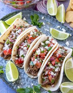 four tacos on a plate with limes and cilantro in the background