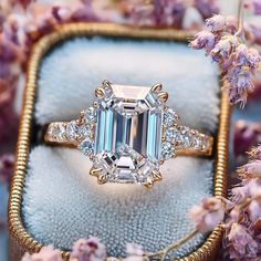 an emerald colored diamond ring sits in a box surrounded by purple flowers and lavenders