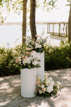 two tall white vases with flowers are on the ground near some trees and water