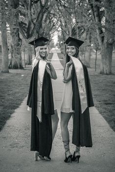 two women in graduation gowns posing for the camera on a path lined with trees