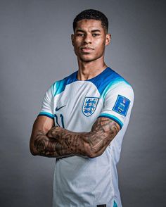 a man with tattoos standing in front of a gray background wearing a white and blue shirt