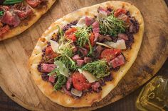 two pizzas with meat and veggies on them sitting on a wooden cutting board