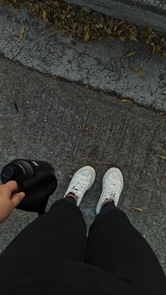 a person with white sneakers and black pants holding a camera on the sidewalk next to some leaves