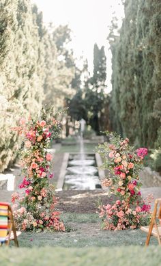 two wooden chairs sitting on top of a grass covered field next to flowers and trees