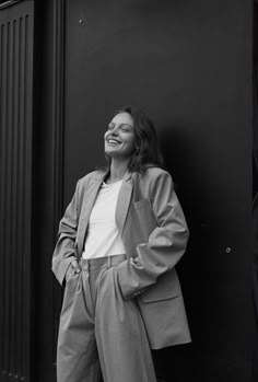 a woman leaning against a wall with her hands in her pockets and smiling at the camera