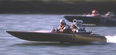 two people in a speed boat on the water with other boats behind them and one person wearing a helmet