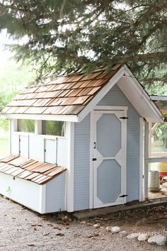 a small blue and white shed with a wooden roof next to a large pine tree