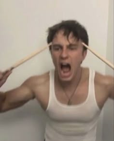 a man holding two wooden sticks over his head and making an angry face while standing in front of a white wall