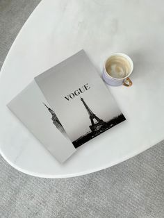 a cup of coffee sitting on top of a white table next to a book with the word vogui