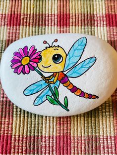 a painted rock with a cartoon insect holding a flower on it's back, sitting on a checkered table cloth