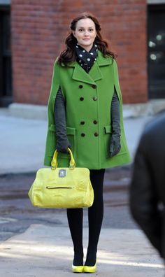 a woman is walking down the street carrying a yellow purse and wearing a green coat