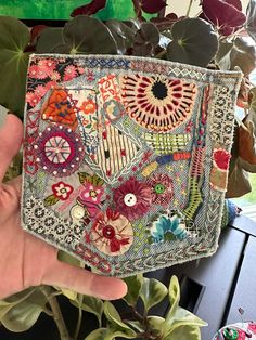 someone is holding up a patchwork purse with buttons and flowers on it in front of some plants