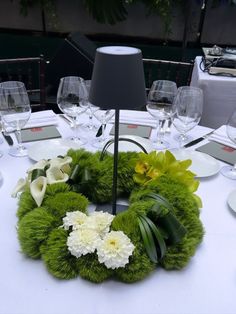 a green wreath with white flowers and greenery sits on a table in front of wine glasses