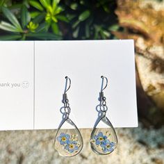 a pair of earrings with blue flowers on them sitting in front of a thank card